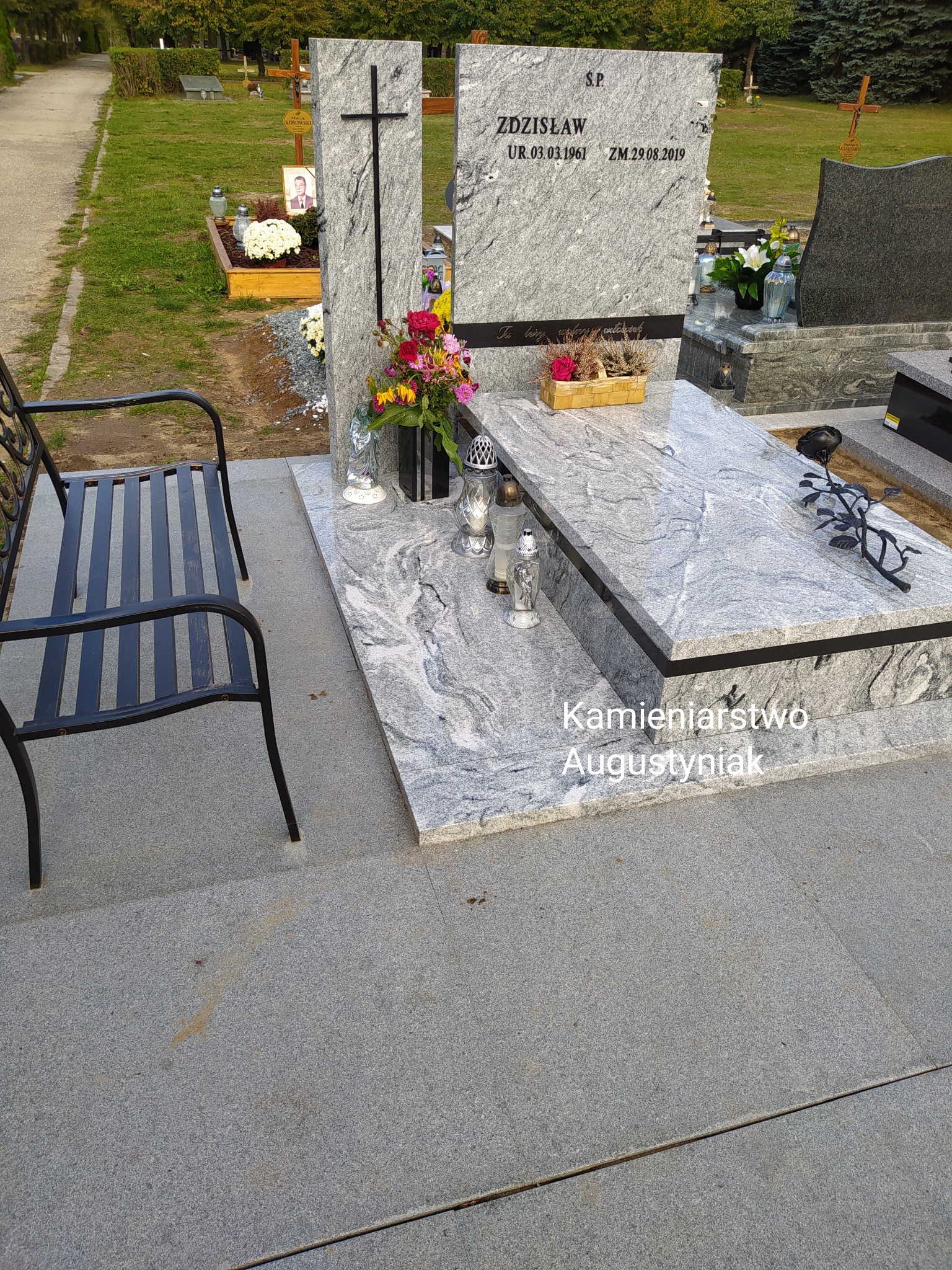 Jasnoszary nagrobek z granitu z czarnym krzyżem i wyrytymi danymi osoby zmarłej, ozdobiony kwiatami i lampionami, obok metalowa ławka, na cmentarzu.