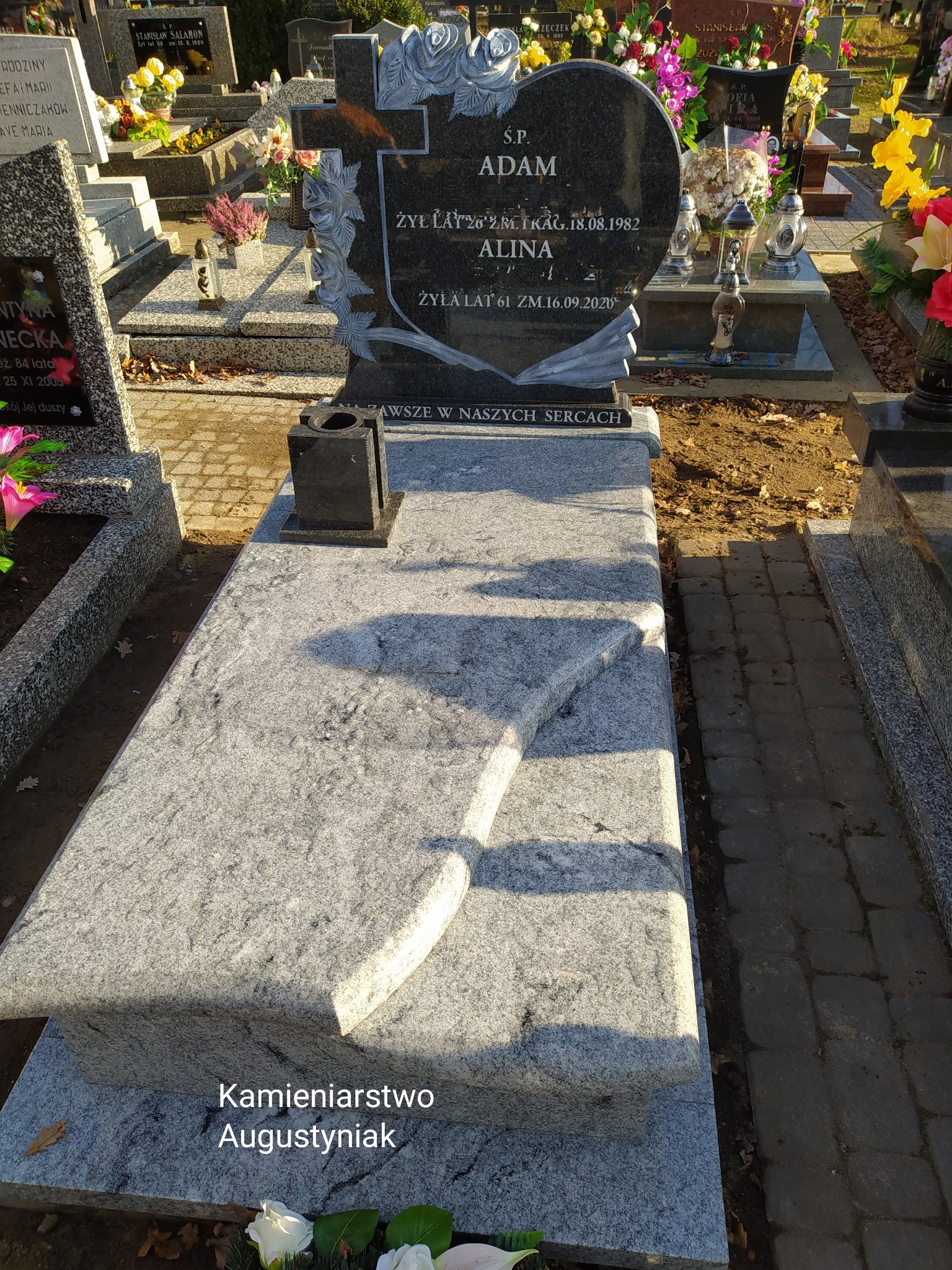 Granitowy nagrobek z krzyżem i sercem, inskrypcje 'S.P. Adam, Alina', kwiatowe dekoracje, jesienne liście na ziemi