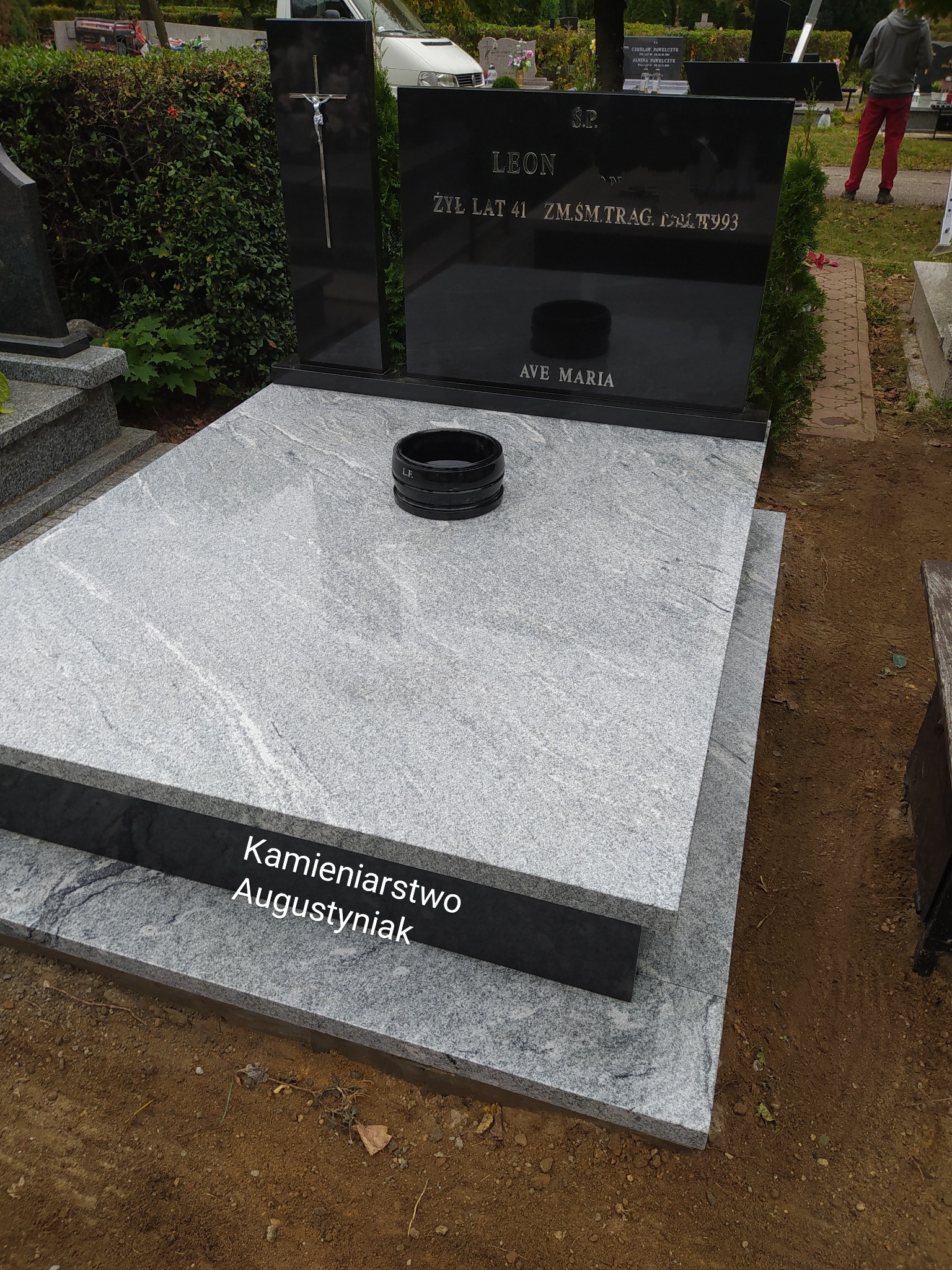 Granitowy nagrobek z czarną tablicą upamiętniającą Leona, z inskrypcją 'Ave Maria' i wazonem na kwiaty, na tle zieleni cmentarza.