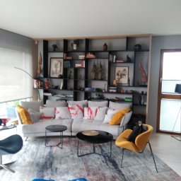 FHU Art-primus - Jasnoszara sofa z wzorzystymi poduszkami, czarne stoliki kawowe i żółty fotel na tle regału z dekoracjami w salonie. Na pierwszym planie niebieska torba.