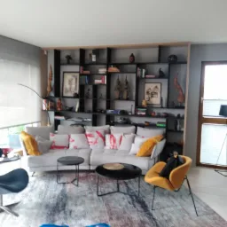 Jasnoszara sofa z wzorzystymi poduszkami, czarne stoliki kawowe i żółty fotel na tle regału z dekoracjami w salonie. Na pierwszym planie niebieska torba.