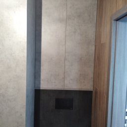 FHU Art-primus - Szare szafki łazienkowe z panelami imitującymi beton, zamontowane nad czarną zabudową WC, obok drewnopodobnych płytek.