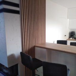 FHU Art-primus - Drewniany blat barowy z wysokimi krzesłami na tle minimalistycznej kuchni z drewnianą ścianką działową z listewek i kominkiem z szarego betonu.