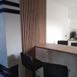 Drewniany blat barowy z wysokimi krzesłami na tle minimalistycznej kuchni z drewnianą ścianką działową z listewek i kominkiem z szarego betonu.