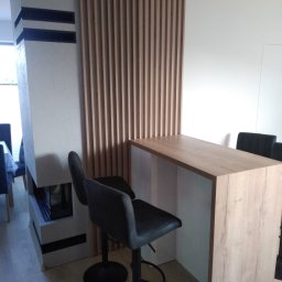 FHU Art-primus - Kuchenny barek z blatem o fakturze jasnego drewna, wsparty na białej podstawie, z dwoma czarnymi hokerami, na tle drewnianej ścianki działowej z pionowych listew i kominka.