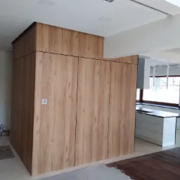 Wysoka szafa wnękowa z drewnopodobnymi frontami w jasnej kuchni, widok na białą zabudowę kuchenną i okno, podłoga częściowo zabezpieczona folią malarską.