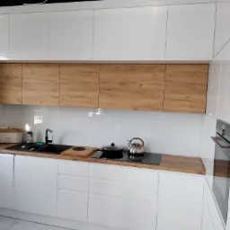 Nowoczesna kuchnia z białymi, lakierowanymi szafkami i drewnianymi frontami górnych szafek, czarny zlew i płyta indukcyjna na blacie z drewnianym wykończeniem, piekarnik w zabudowie.