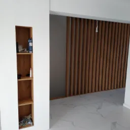 Biała ściana z wnęką na p&oacute;łki z drewnianym wnętrzem, widoczna zawartość p&oacute;łek, obok drewniana ażurowa ścianka działowa, marmurowa podłoga.