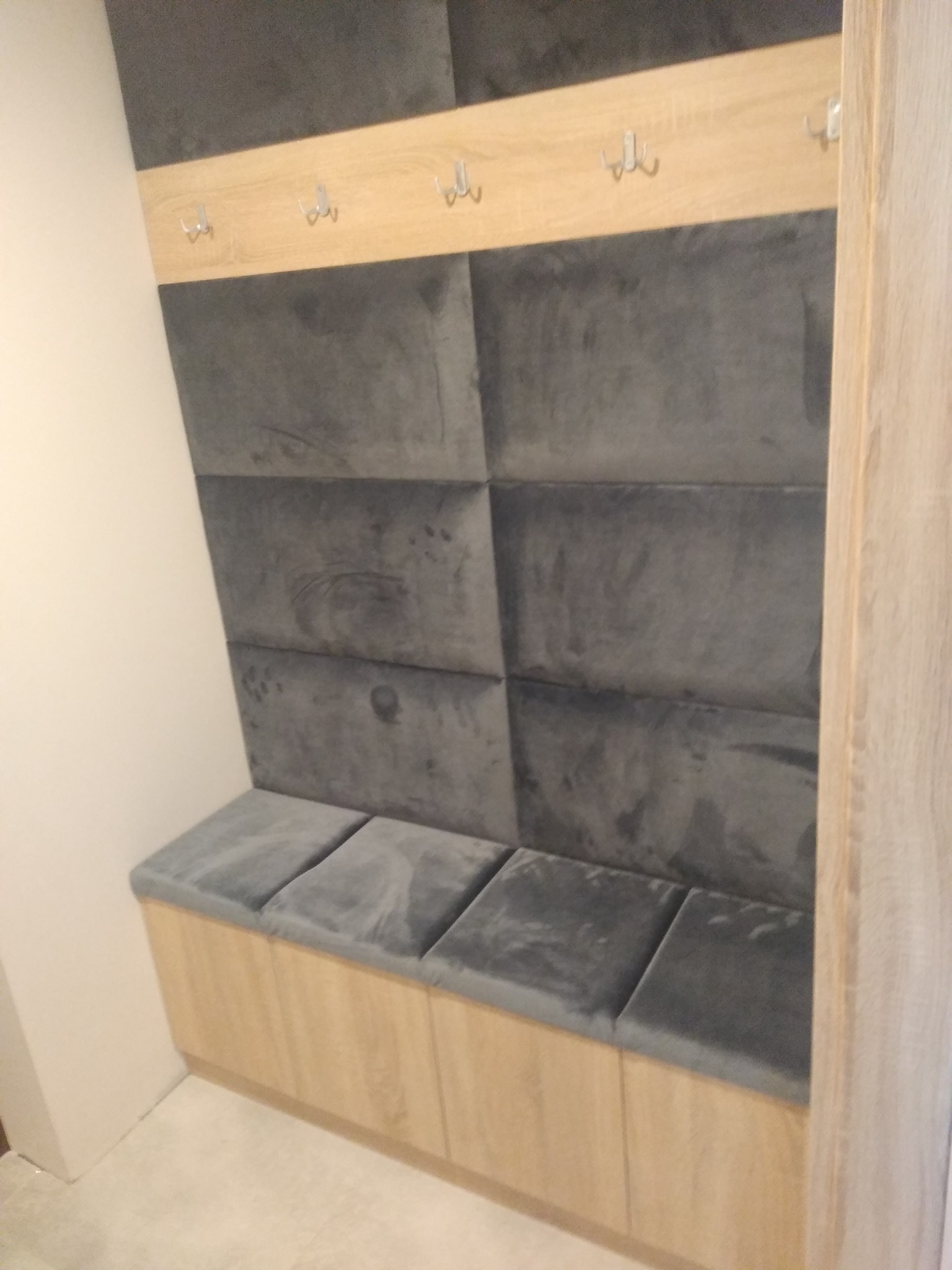 Szafa wnękowa z siedziskiem wykończonym szarymi panelami tapicerowanymi, drewniany panel z wieszakami na ubrania.