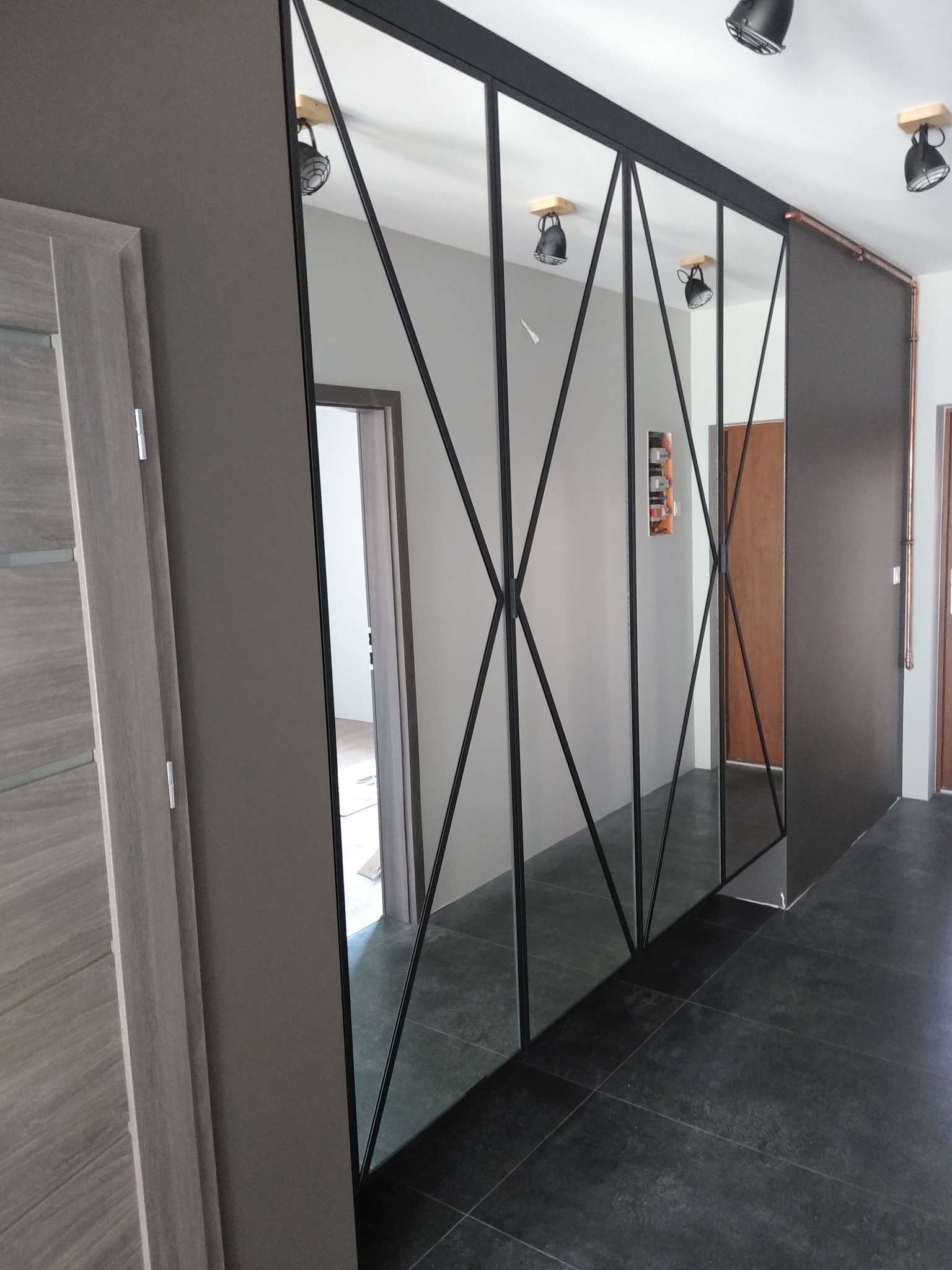 Nowoczesna ściana działowa z lustrami w czarnych ramach, tworząca geometryczny wzór w korytarzu z ciemną podłogą i jasnymi ścianami.