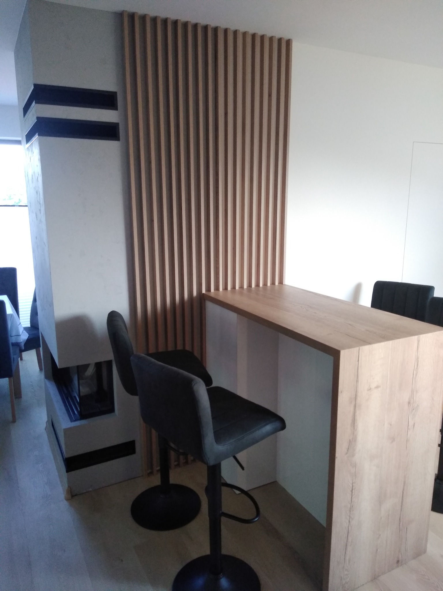 Kuchenny barek z blatem o fakturze jasnego drewna, wsparty na białej podstawie, z dwoma czarnymi hokerami, na tle drewnianej ścianki działowej z pionowych listew i kominka.