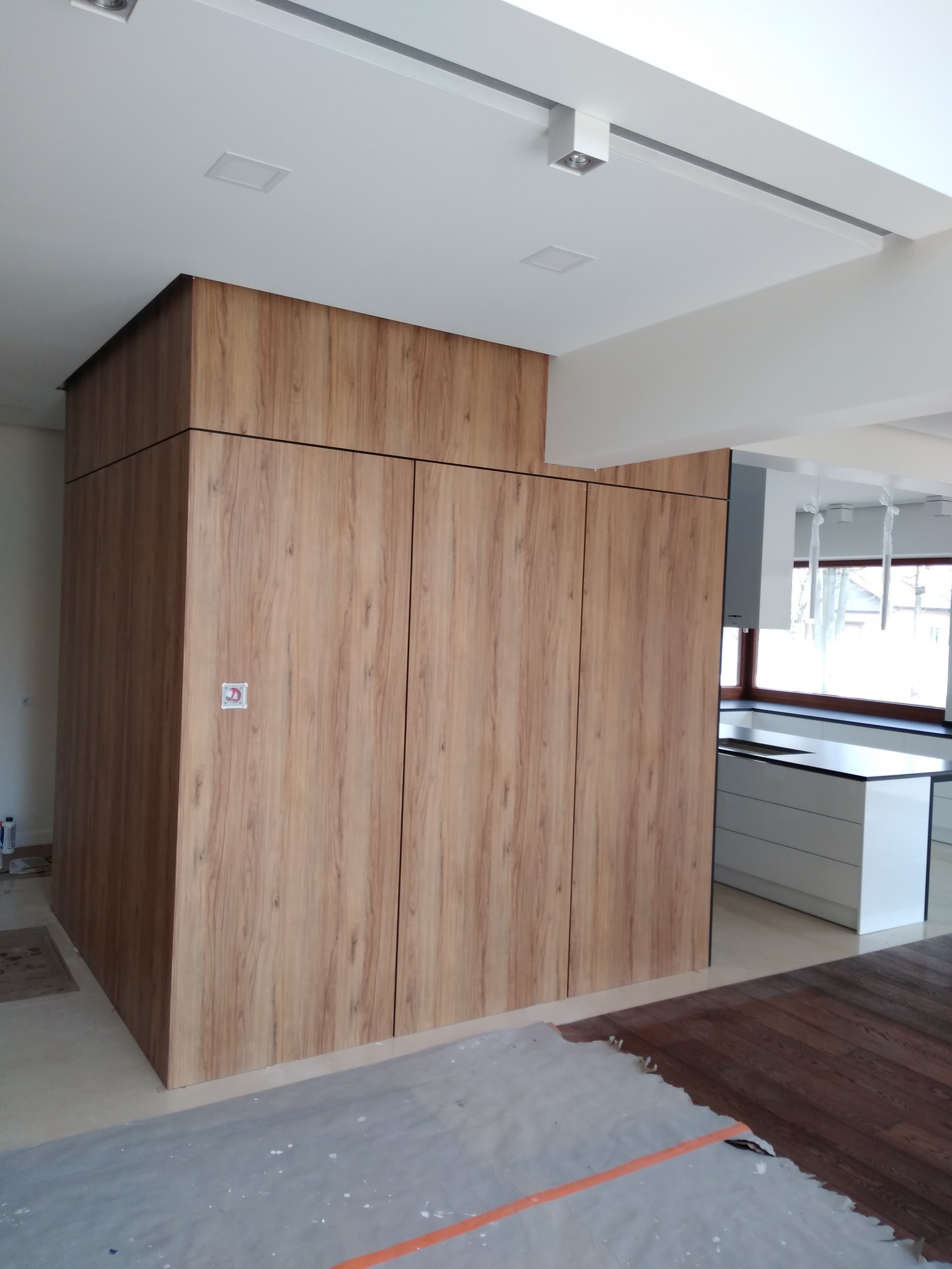 Wysoka szafa wnękowa z drewnopodobnymi frontami w jasnej kuchni, widok na białą zabudowę kuchenną i okno, podłoga częściowo zabezpieczona folią malarską.