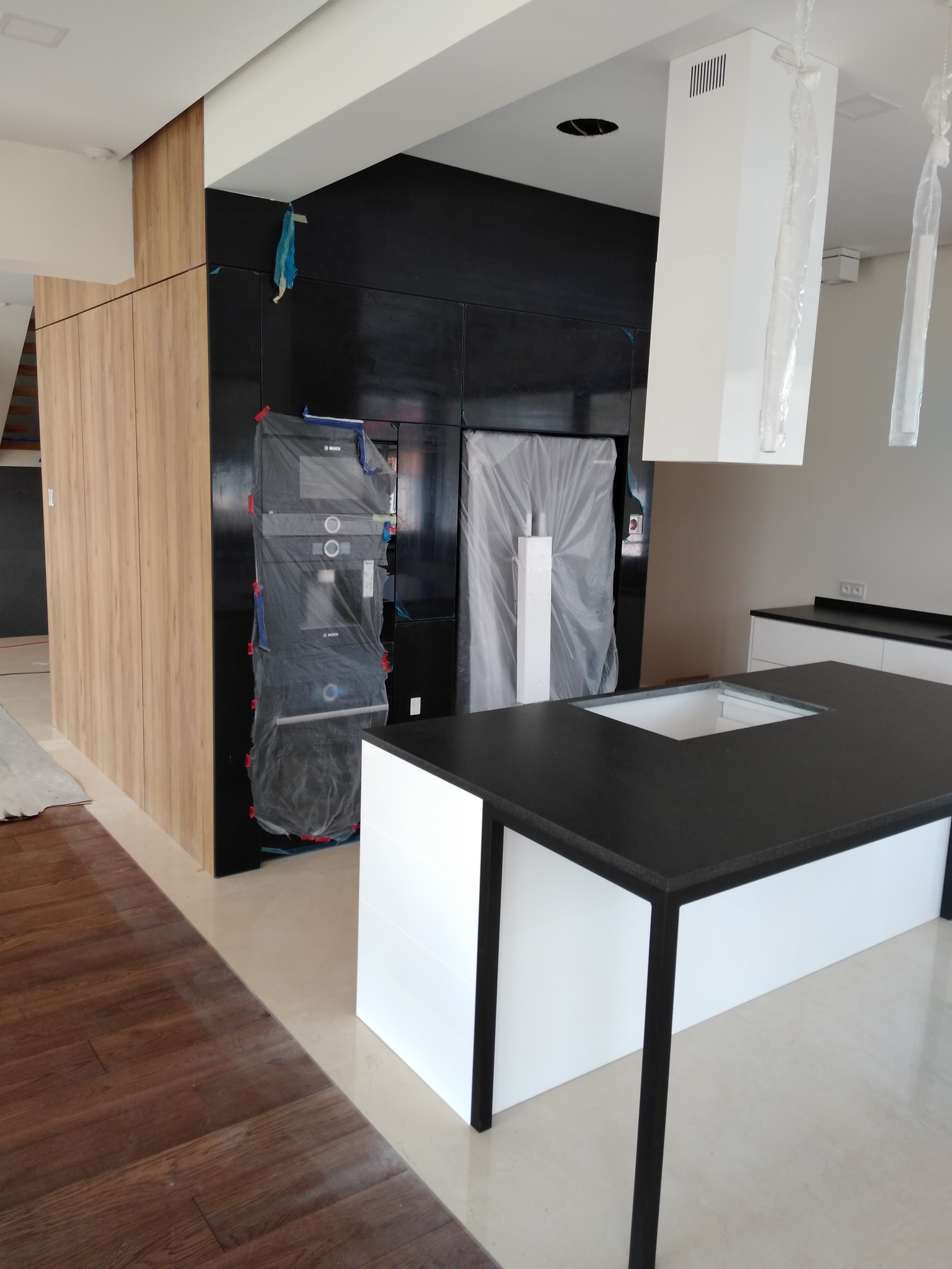 Częściowo zrealizowana kuchnia z wyspą, czarnym blatem i sprzętami AGD pod folią ochronną, drewniane fronty szafek po lewej, minimalistyczny design.