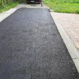 Świeżo położona, czarna nawierzchnia asfaltowa przylega do chodnika z kolorowej kostki brukowej, w tle widoczny fragment zaparkowanego samochodu.