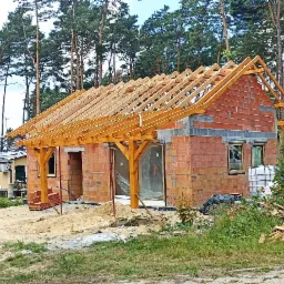 Surowy stan budynku jednorodzinnego z drewnianą konstrukcją dachu w trakcie budowy, widok z zewnątrz, z stertą drewna i piasku na posesji.