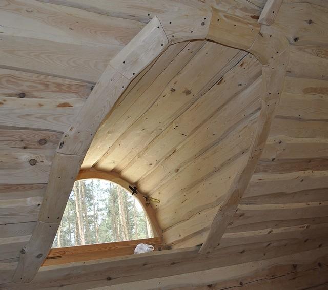 Wnętrze poddasza wykończonego jasnym drewnem z oknem dachowym w kształcie łuku, widok na las przez okno, widoczne łączenia drewnianych elementów.