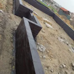 Świeżo zaizolowane fundamenty domu w trakcie budowy, widoczne ślady po pędzlu na czarnej powłoce hydroizolacyjnej, ziemia wok&oacute;ł wykopu, w tle domy jednorodzinne.