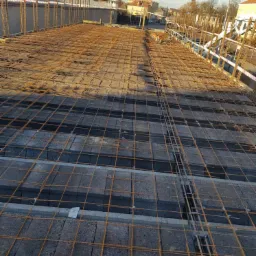 Zbrojenie fundamentów z użyciem stalowych prętów i bloczków betonowych, widok z góry na plac budowy w słoneczny dzień.