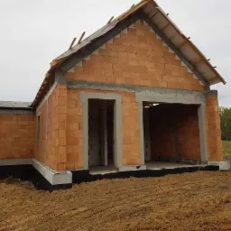 Surowy stan domu murowanego z pustaków ceramicznych, widoczne fundamenty z izolacją, brak okien i drzwi, przygotowany dach bez pokrycia, teren wokół wyrównany.