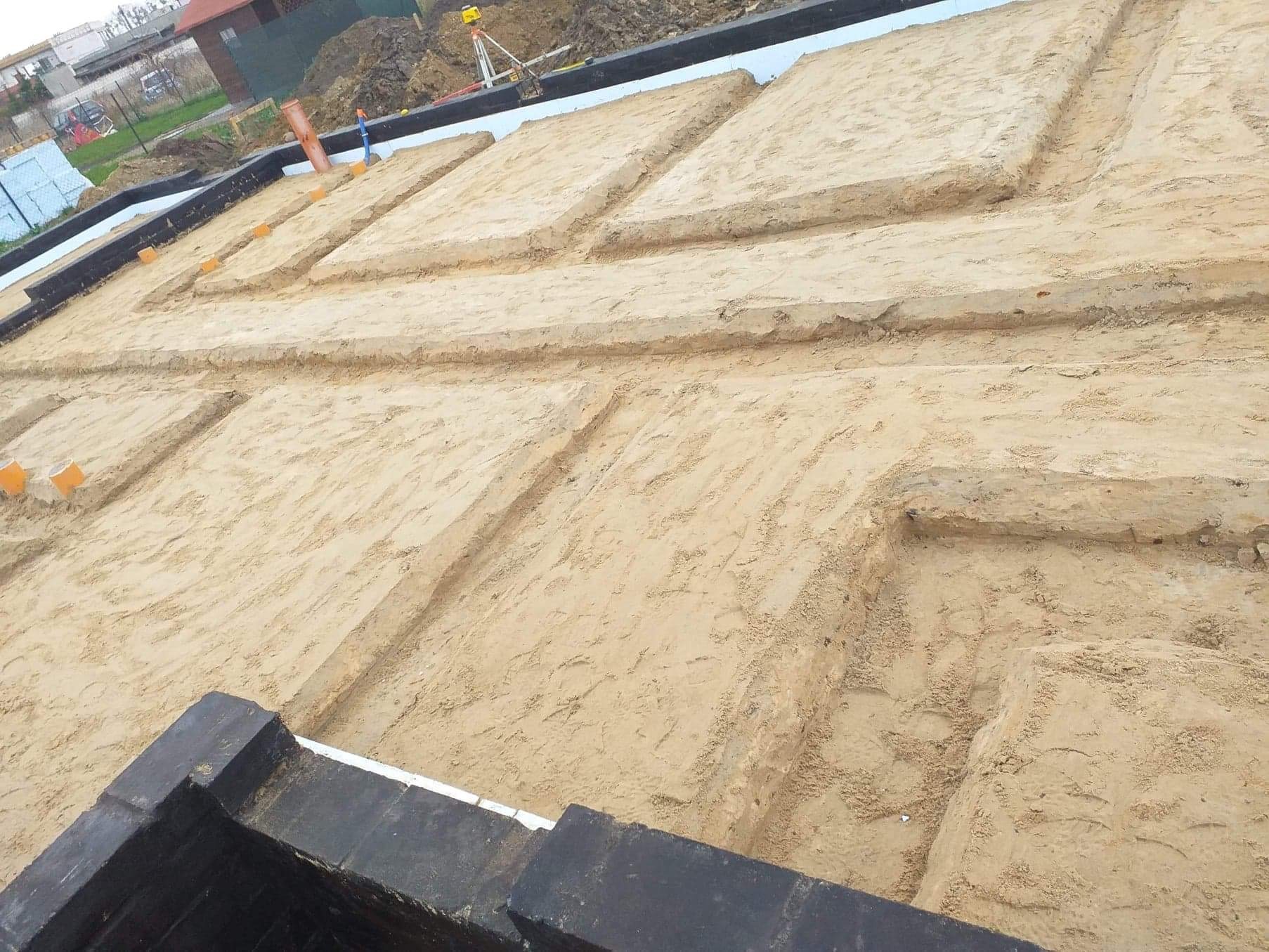 Przygotowanie wykopów pod ławy fundamentowe, widoczne szalunki i izolacja przeciwwilgociowa, w tle niwelator laserowy.