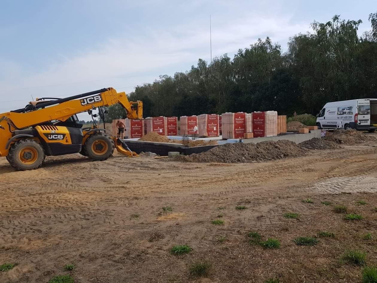 Budowa domu murowanego: żółta ładowarka teleskopowa JCB ustawia cegły ceramiczne na fundamencie z izolacją pionową, w tle biały samochód dostawczy i las.