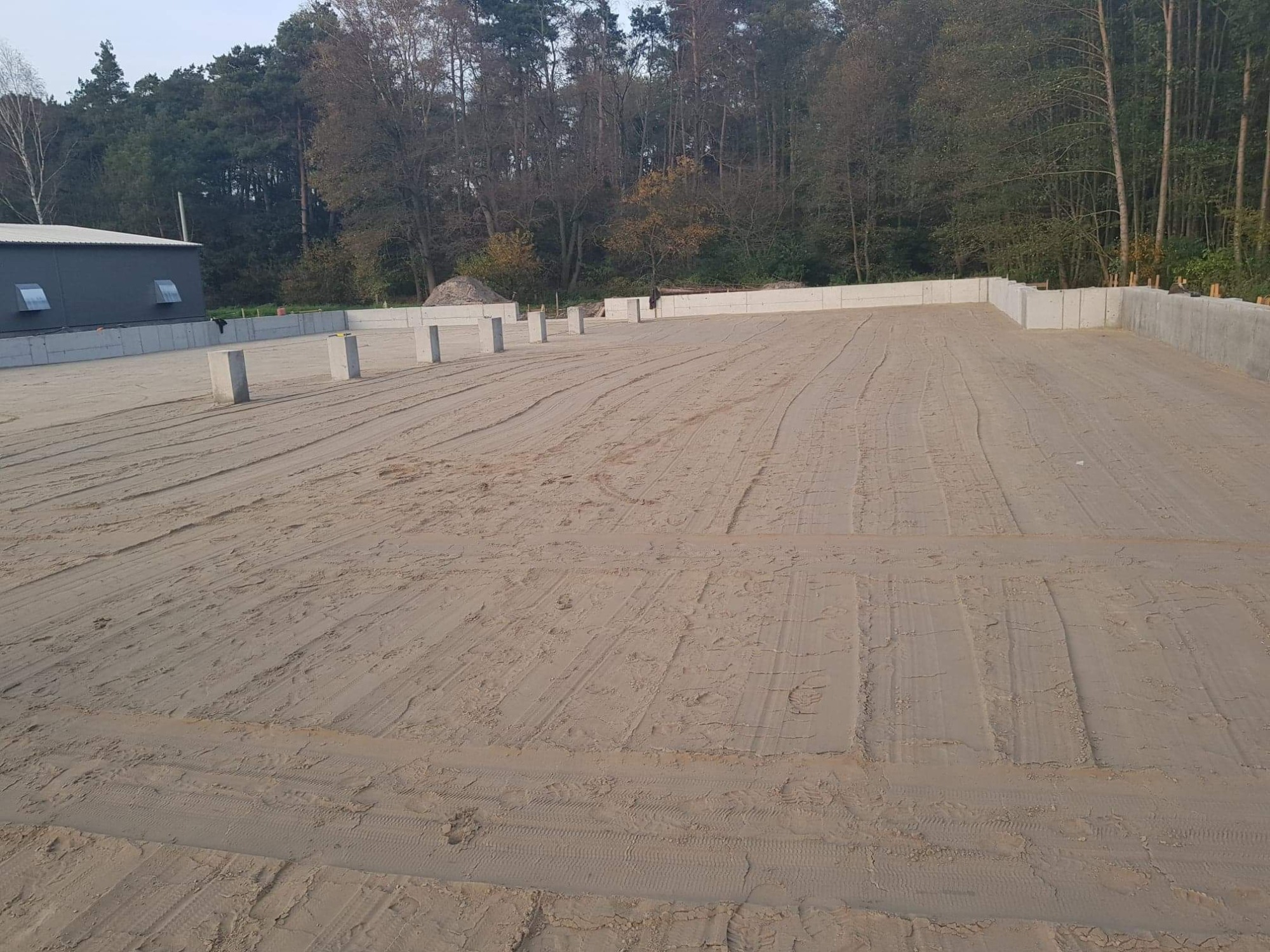 Przygotowany teren pod budowę, widoczne betonowe fundamenty i słupy, ślady opon na piasku, budynek gospodarczy w tle, jesienne drzewa.