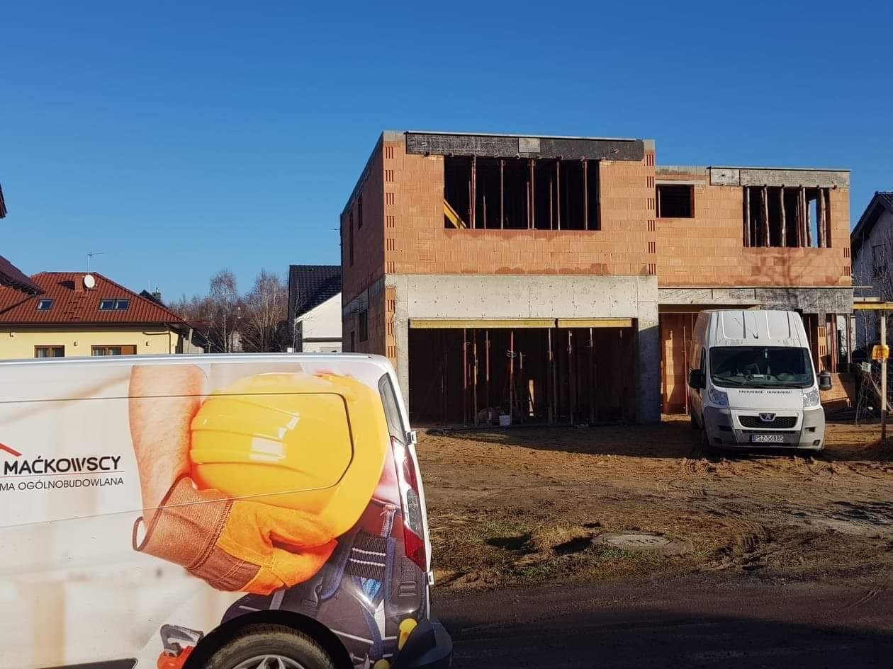 Niedokończony dwukondygnacyjny budynek z czerwonej cegły i szarego betonu, z widoczną konstrukcją garażu, na tle błękitnego nieba, z samochodem dostawczym z logo firmy budowlanej na pierwszym planie.