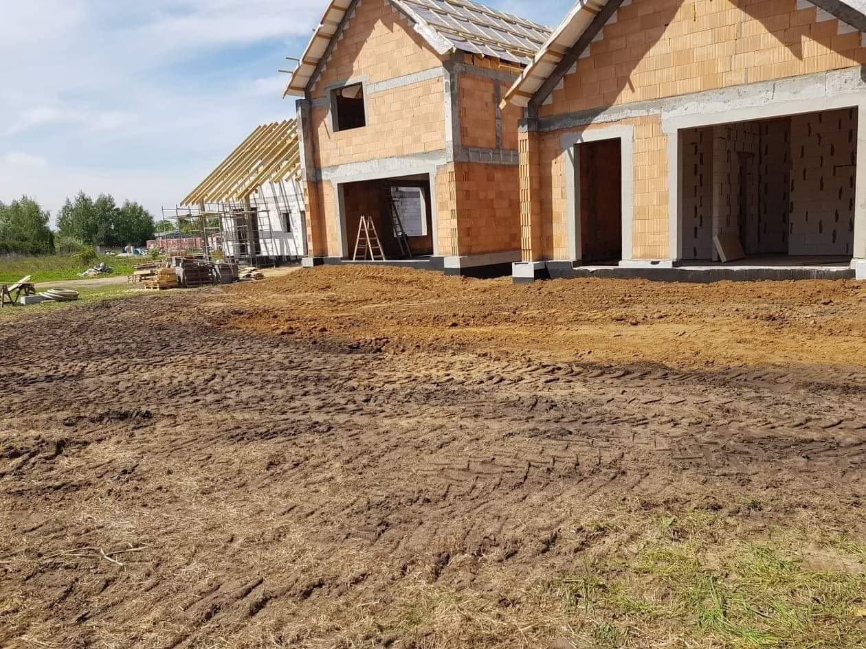 Wznoszenie dwóch budynków jednorodzinnych z czerwonej cegły, widoczne fundamenty, niezadaszona konstrukcja dachu jednego z budynków, teren budowy z wyjeżdżoną ziemią.