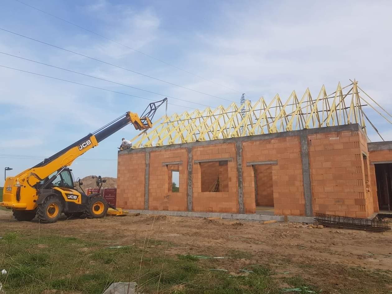 Budowa domu z czerwonej cegły: widoczny stan surowy otwarty, z drewnianą konstrukcją dachu w trakcie montażu, oraz żółta ładowarka teleskopowa JCB z operatorem na górze muru.