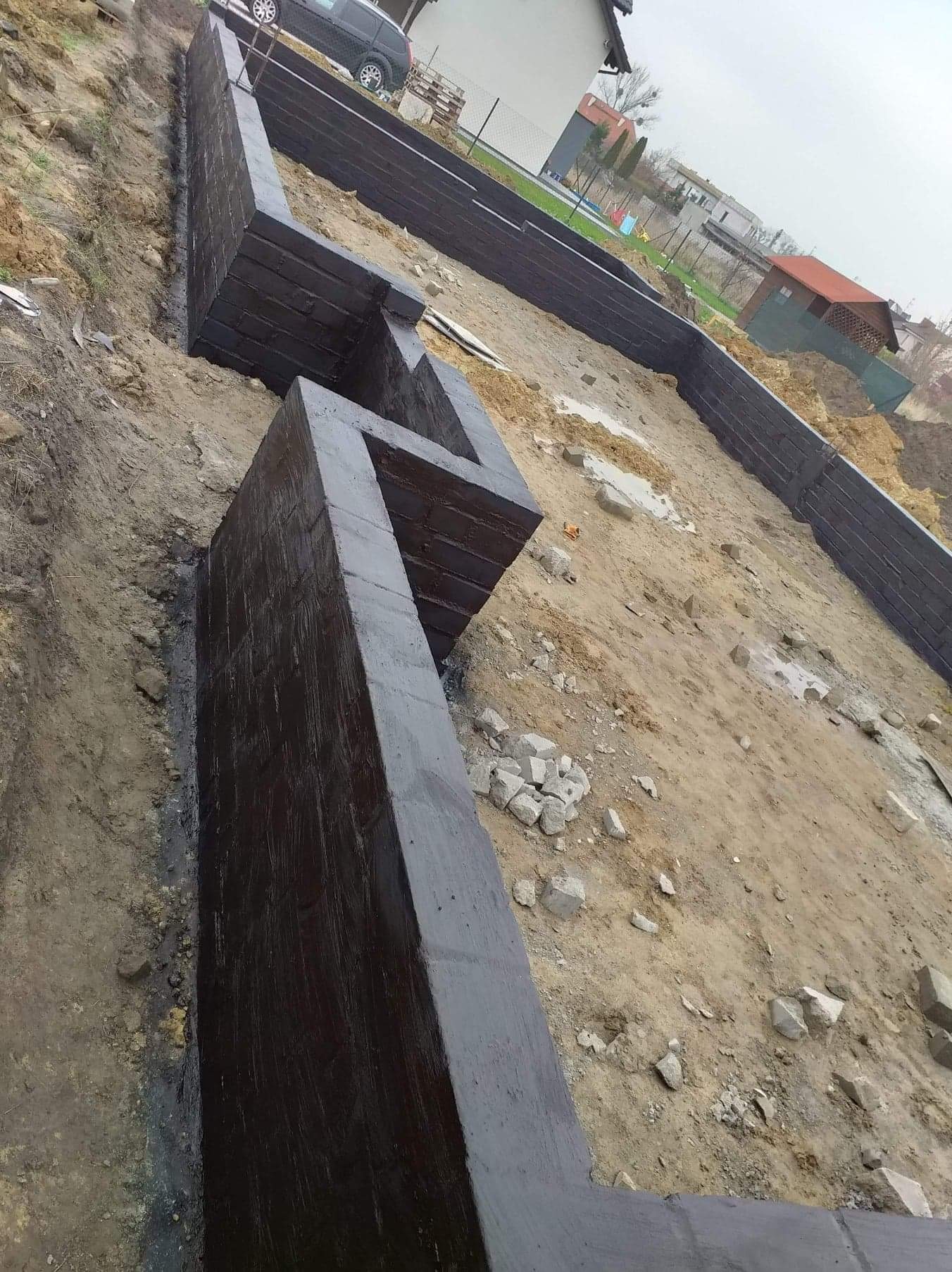 Świeżo zaizolowane fundamenty domu w trakcie budowy, widoczne ślady po pędzlu na czarnej powłoce hydroizolacyjnej, ziemia wokół wykopu, w tle domy jednorodzinne.