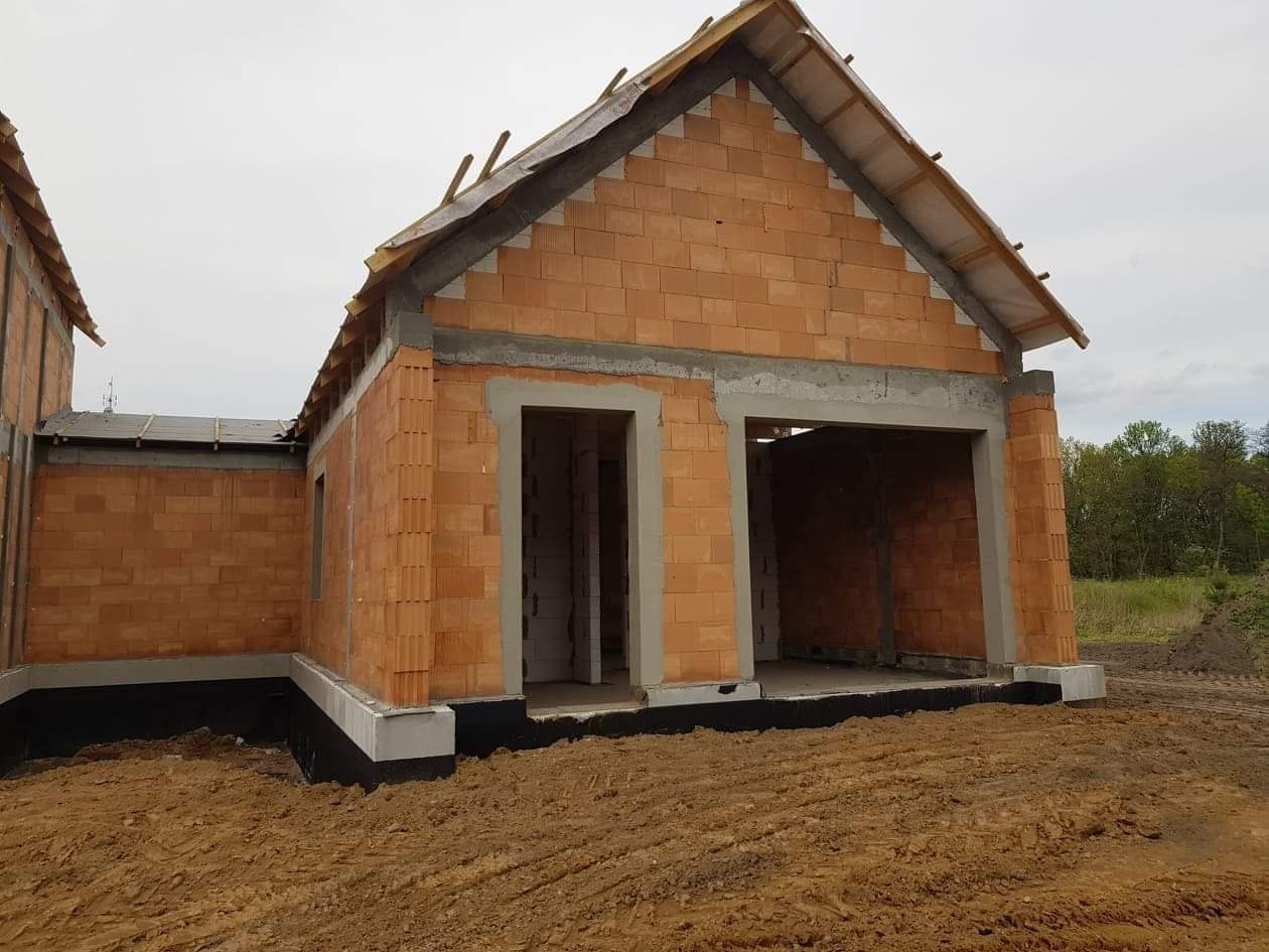 Surowy stan domu murowanego z pustaków ceramicznych, widoczne fundamenty z izolacją, brak okien i drzwi, przygotowany dach bez pokrycia, teren wokół wyrównany.