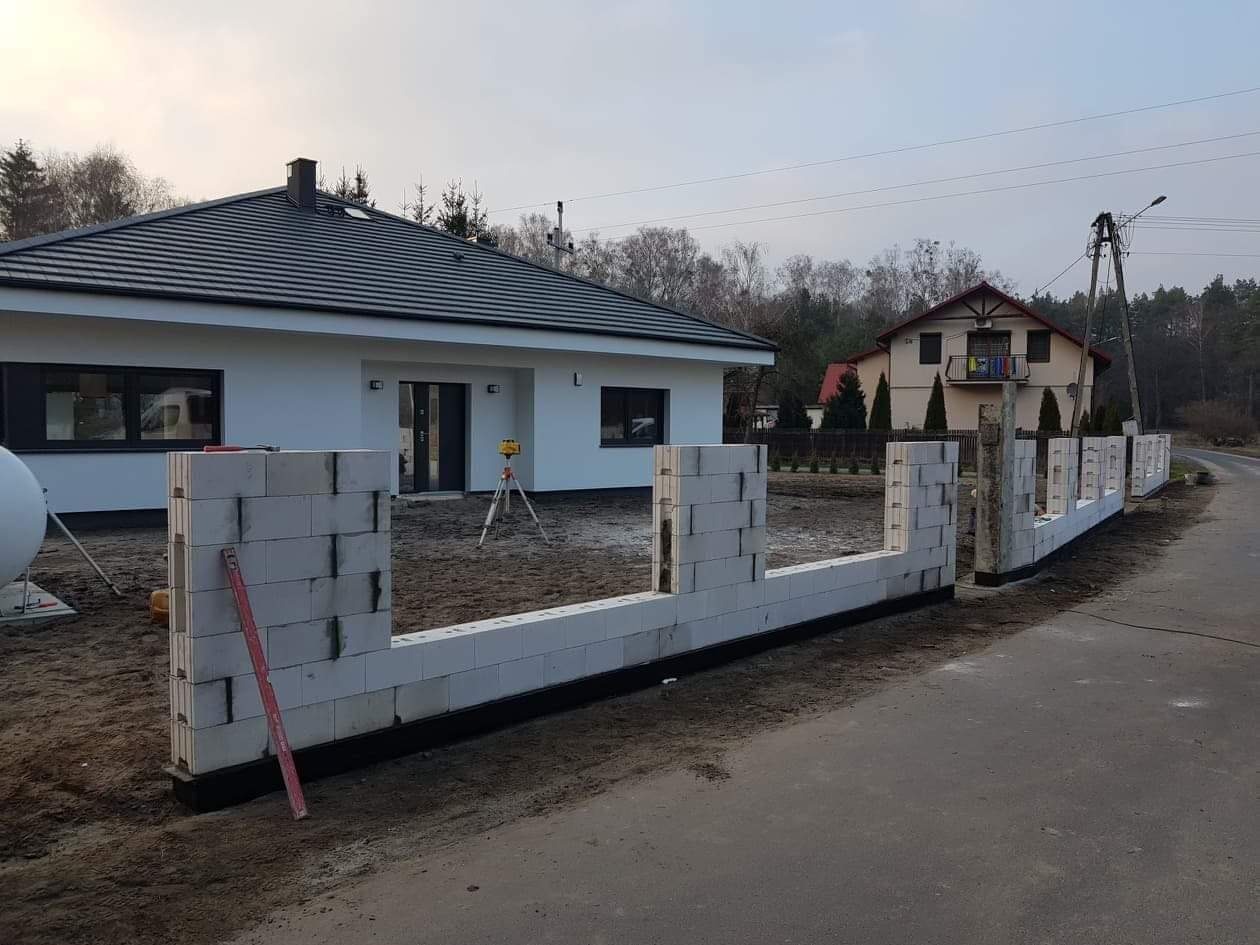 Budowa ogrodzenia z białych bloczków betonowych, widoczny poziomica i niwelator laserowy, w tle dom jednorodzinny z ciemnym dachem.