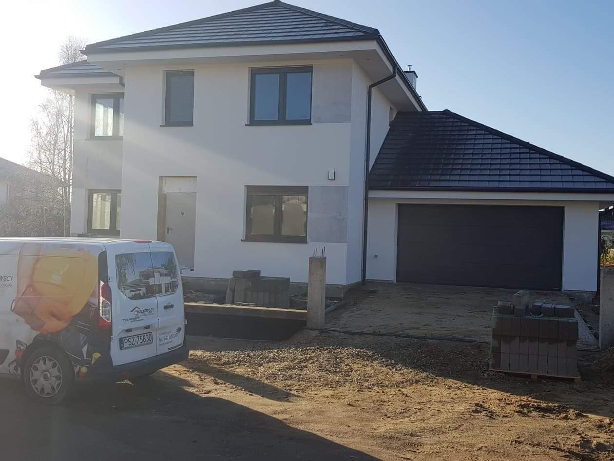 Dwukondygnacyjny budynek w trakcie wykończenia elewacji, widoczna biała fasada, grafitowa dachówka, brama garażowa i zaparkowany samochód dostawczy z logo firmy.