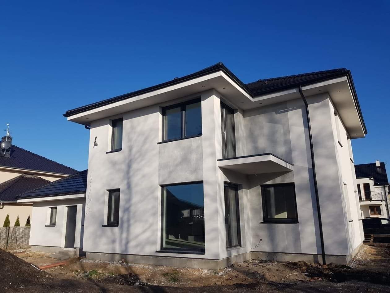 Nowoczesny dom dwukondygnacyjny z szarą elewacją i czarną stolarką okienną, widoczne rynny i fragment dachu sąsiedniego budynku.