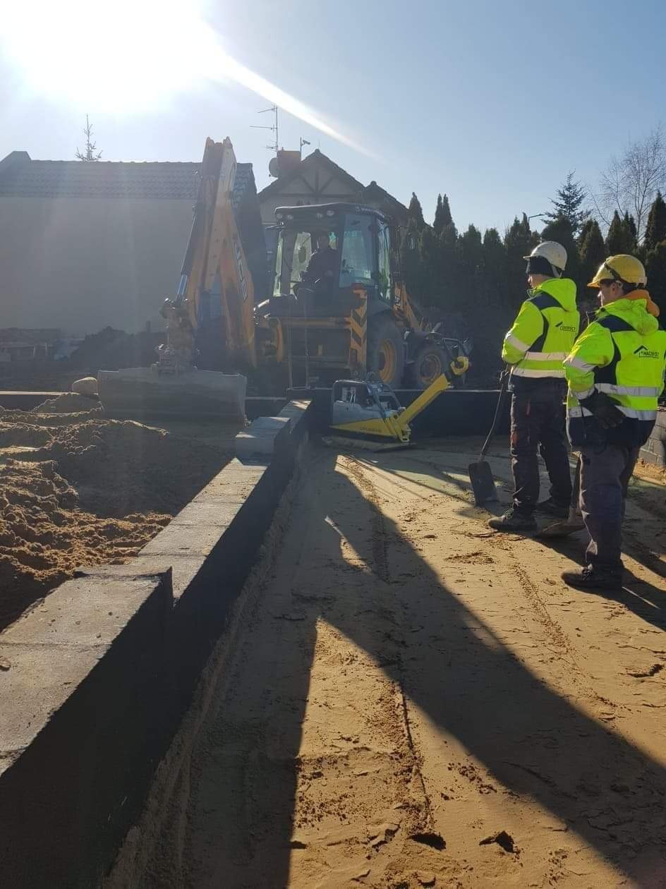 Przygotowanie terenu pod fundamenty: wykop, ubijarka, pracownicy w odblaskowych kurtkach i koparka w tle.