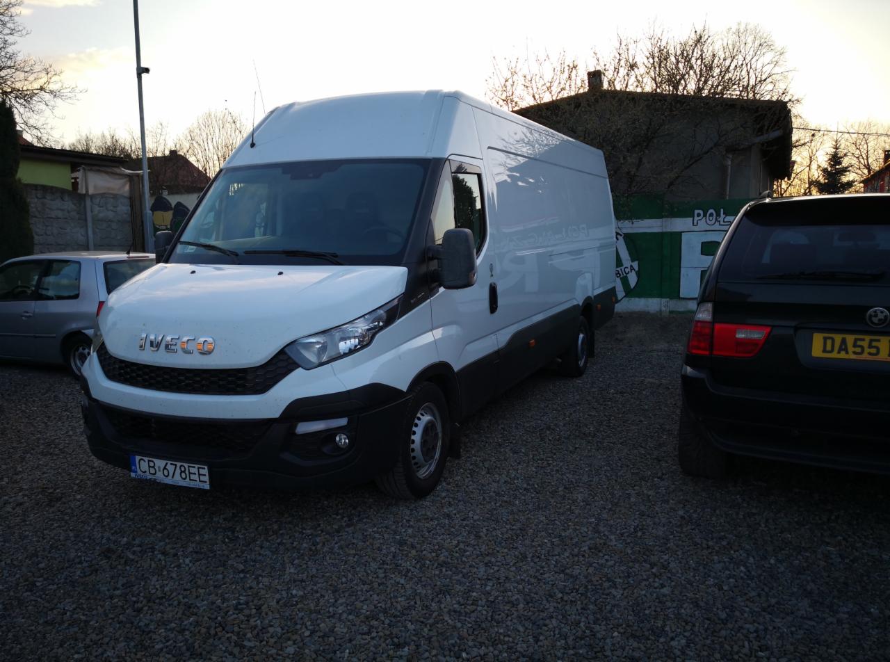 Biały furgon Iveco na żwirowym parkingu, obok zaparkowany czarny SUV BMW, w tle widoczny mur i drzewa.