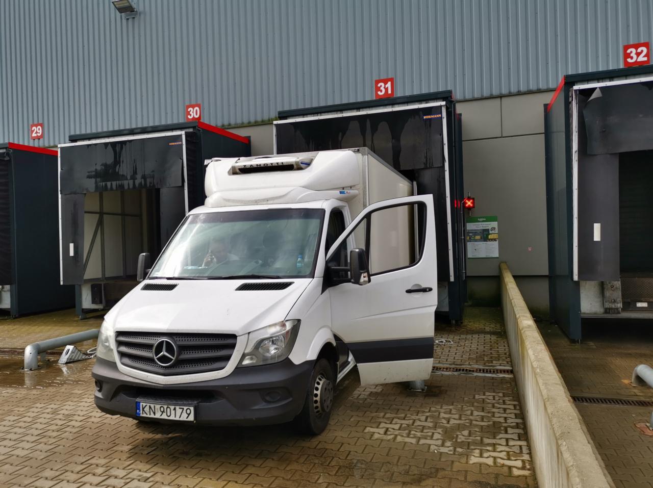 Biały bus chłodnia Mercedes z otwartymi drzwiami stoi przed rampą załadunkową z numerem 31, widoczna tablica rejestracyjna KN 90179.