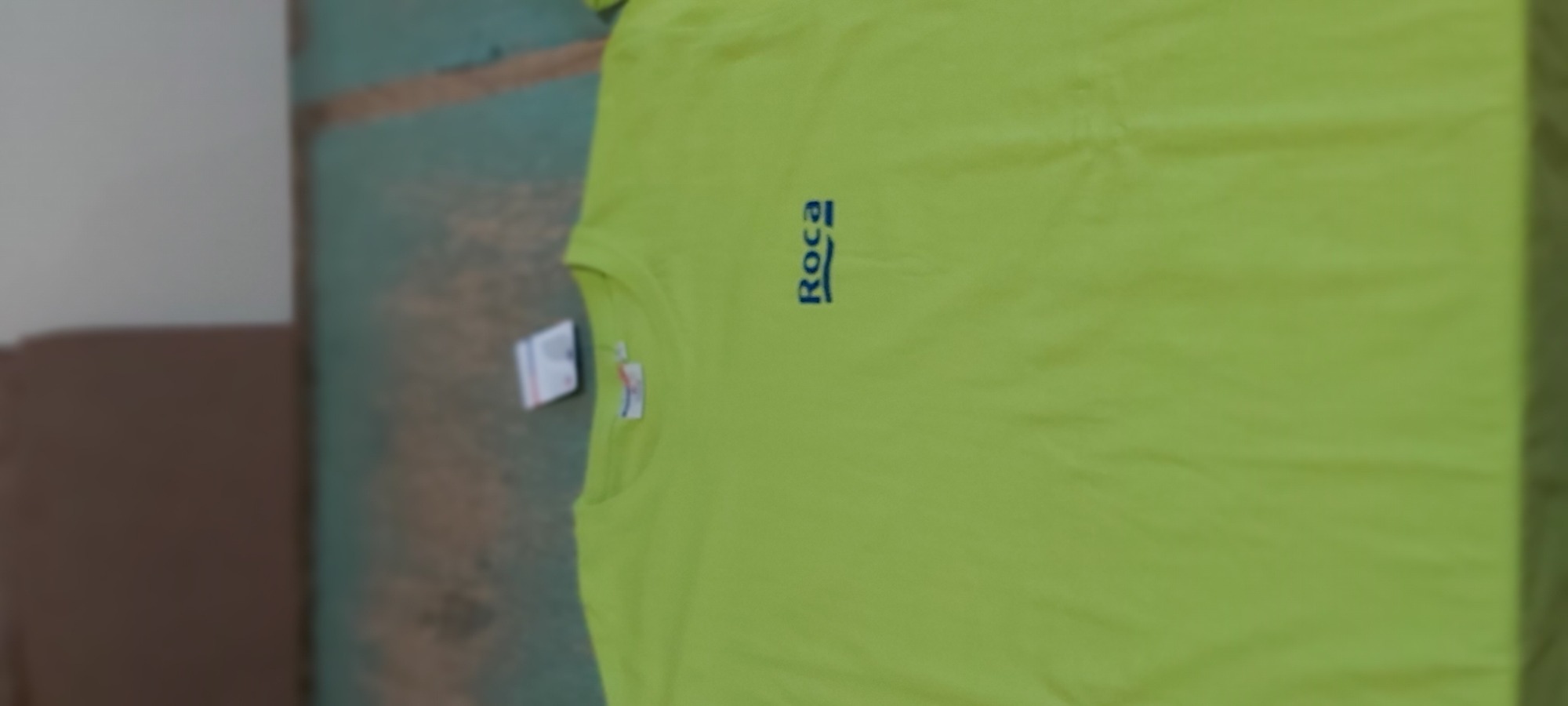 Jasnozielona koszulka z krótkim rękawem i niebieskim logo Roca, leżąca na nierównym tle z etykietą