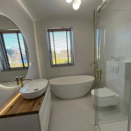 KABUD Kamil Karkowski - Nowoczesna łazienka z owalną wanną wolnostojącą, umywalką nablatową na drewnianym blacie, kabiną prysznicową ze złotymi elementami i heksagonalnymi płytkami na ścianach.