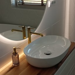 KABUD Kamil Karkowski - Biała, owalna umywalka nablatowa z złotą armaturą i dozownikiem mydła na drewnianym blacie z podświetleniem LED, na tle heksagonalnych płytek i okrągłego lustra z podświetleniem.