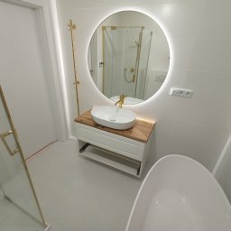 KABUD Kamil Karkowski - Nowoczesna łazienka w bieli z elementami złota: okrągłe lustro z podświetleniem LED, umywalka nablatowa na drewnianym blacie, kabina prysznicowa ze złotymi detalami i biała wanna wolnostojąca.