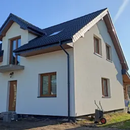 Dwupiętrowy dom po ociepleniu elewacji w słoneczny dzień. Widoczne okna z drewnianymi ramami, czarny dach i rynny. Na trawniku stoi taczka i wiaderka z farbą.