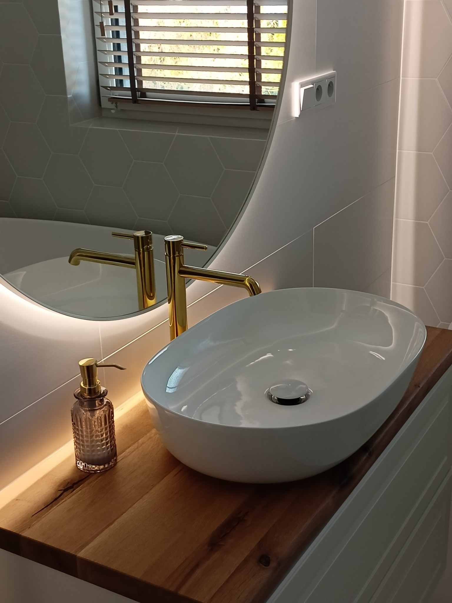 Biała, owalna umywalka nablatowa z złotą armaturą i dozownikiem mydła na drewnianym blacie z podświetleniem LED, na tle heksagonalnych płytek i okrągłego lustra z podświetleniem.