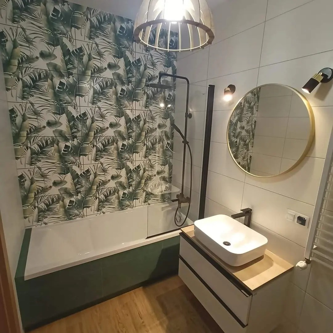 Nowoczesna łazienka z wanną obudowaną zielonymi płytkami, ściana z motywem liści palm, okrągłe lustro w złotej ramie i biała umywalka nablatowa.