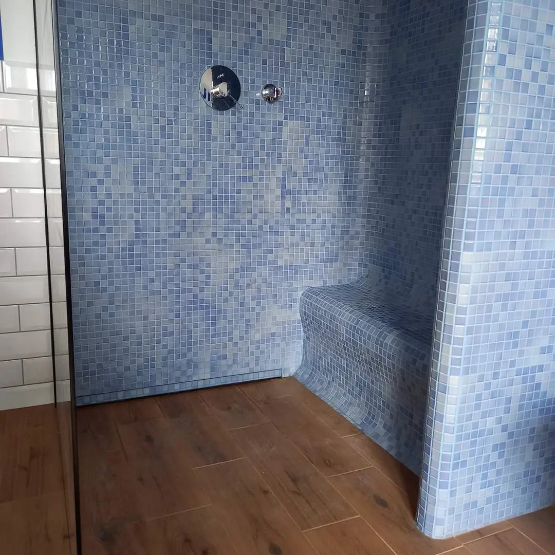 Niebieska mozaika w kabinie prysznicowej z siedziskiem, drewnopodobne płytki podłogowe, chromowana bateria natryskowa.
