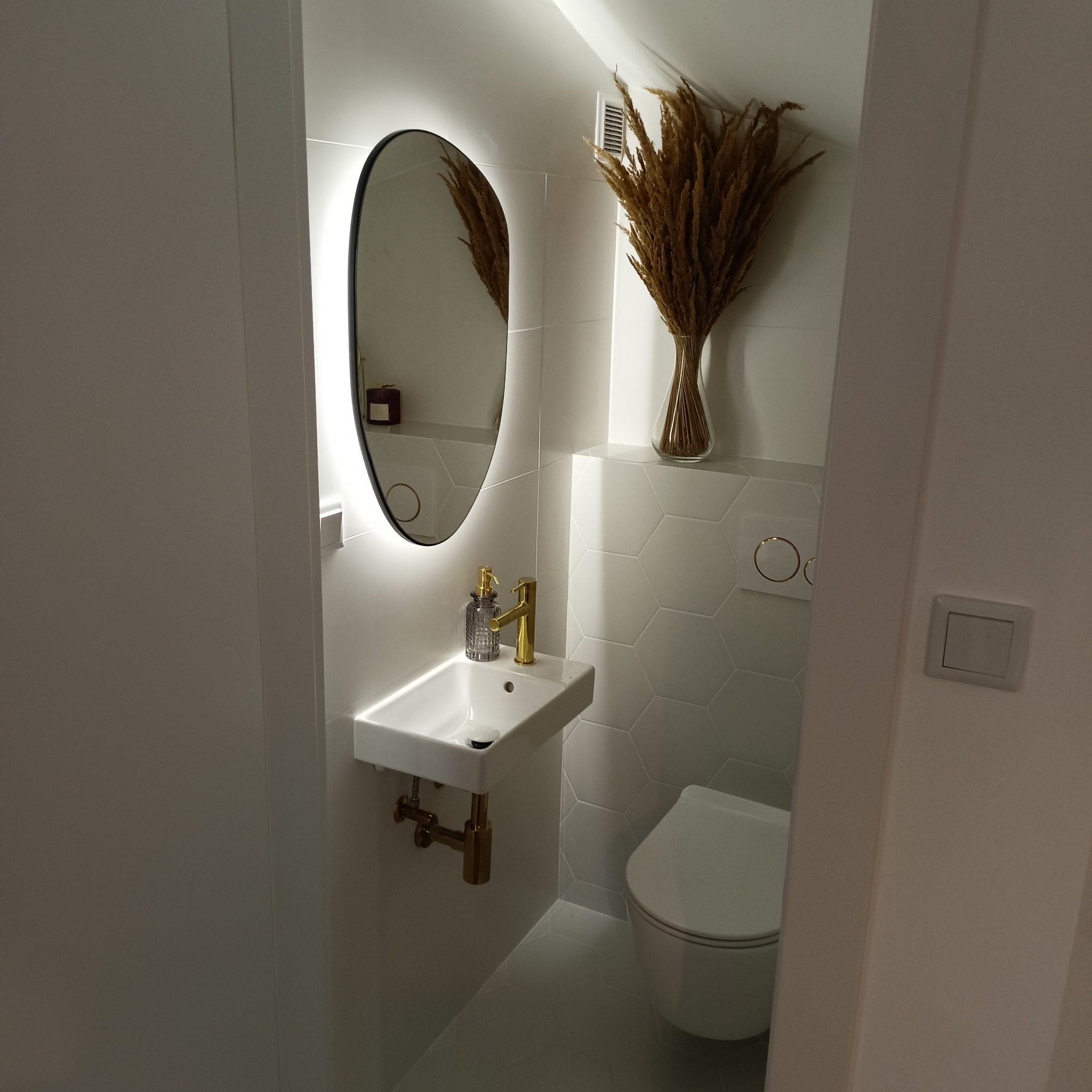 Nowoczesna toaleta dla gości z owalnym lustrem z podświetleniem LED, białą umywalką z baterią w kolorze złotym, heksagonalnymi płytkami i dekoracyjną trawą pampasową w wazonie.