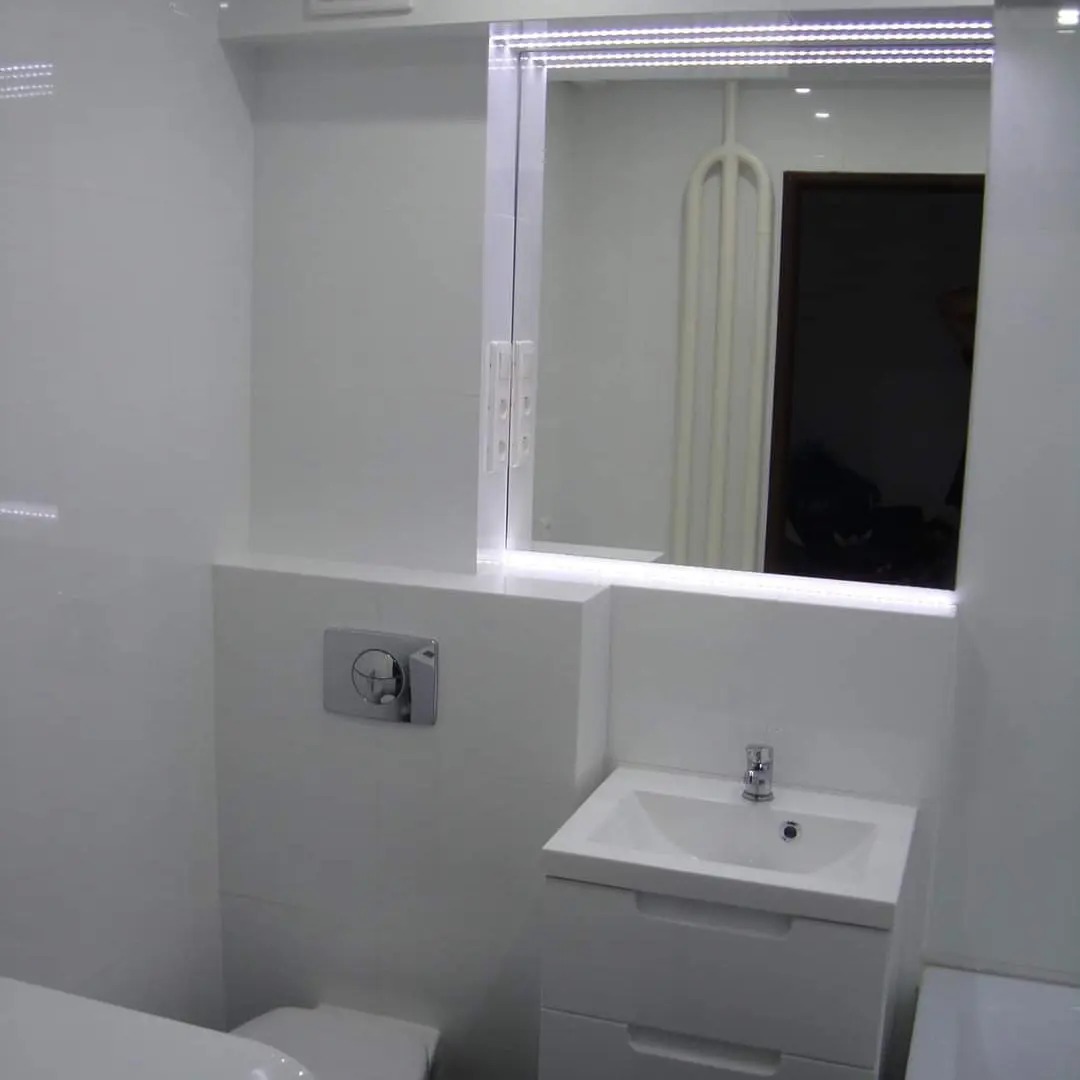 Biała łazienka z podświetlanym lustrem, umywalką z szafką i chromowanym przyciskiem spłuczki, widoczne odbicie w lustrze.