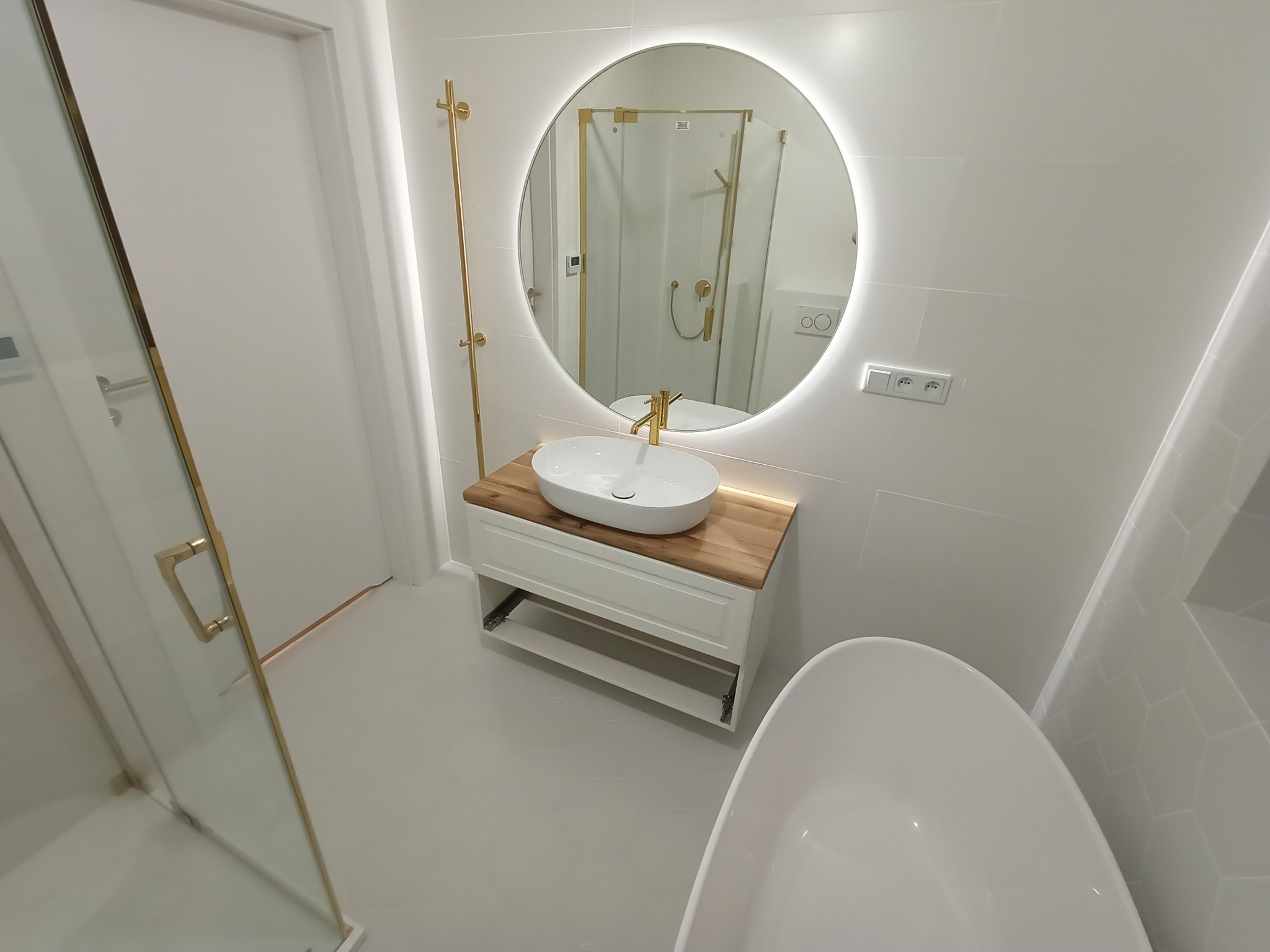 Nowoczesna łazienka w bieli z elementami złota: okrągłe lustro z podświetleniem LED, umywalka nablatowa na drewnianym blacie, kabina prysznicowa ze złotymi detalami i biała wanna wolnostojąca.