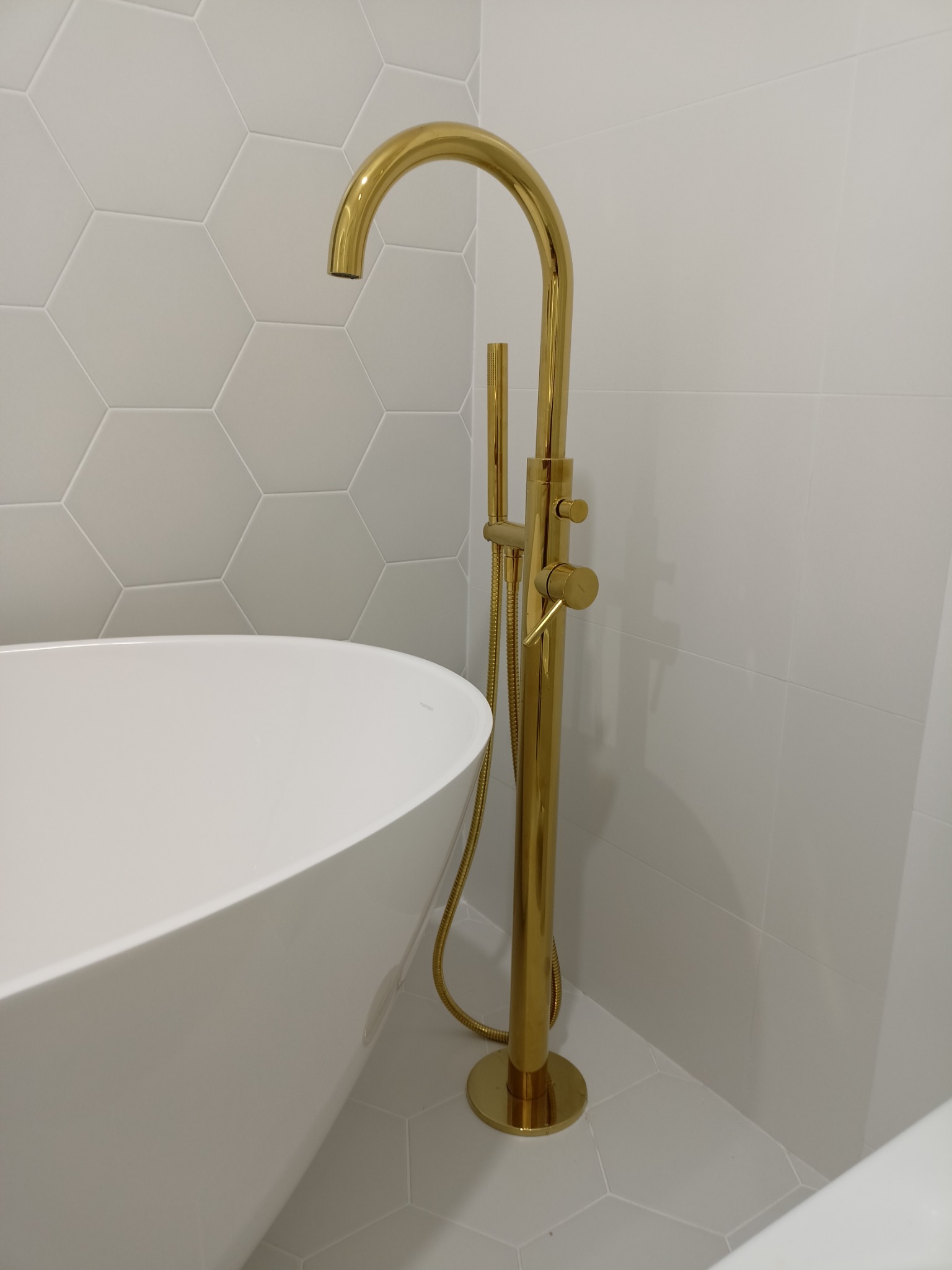 Wolnostojąca, złota bateria wannowa obok białej, owalnej wanny, na tle białych, heksagonalnych płytek ściennych i kwadratowych płytek podłogowych. Widoczny fragment wanny.