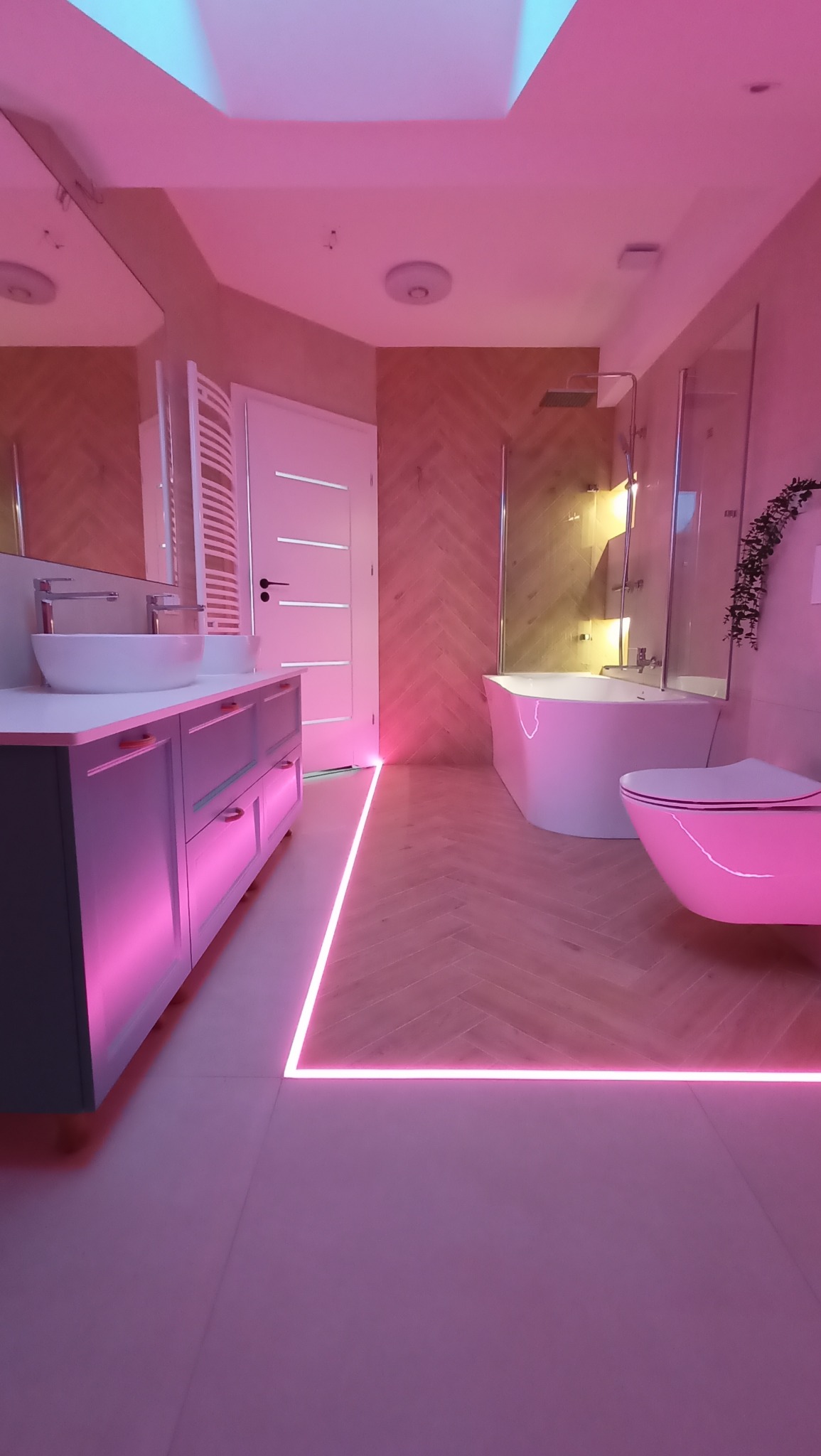 Nowoczesna łazienka z podświetleniem LED w kolorze różowym, umywalki nablatowe, biała wanna i toaleta, drewnopodobna podłoga ułożona w jodełkę.